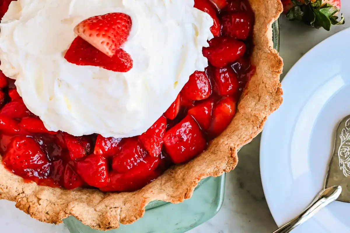 A freshly baked strawberry pie with a golden, flaky sourdough crust, filled with vibrant, juicy strawberries coated in a glossy homemade glaze. The rustic, whole wheat crust adds a rich, nutty flavor that perfectly complements the sweet and tangy strawberry filling. Served in a classic glass pie dish, this delicious dessert is ready to be enjoyed with a fork and plate for the perfect homemade treat.