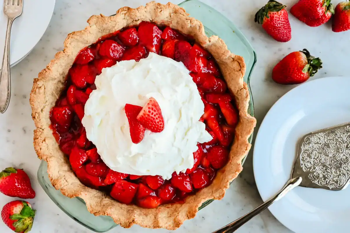 A freshly baked strawberry pie with a golden, flaky sourdough crust, filled with vibrant, juicy strawberries coated in a glossy homemade glaze. The rustic, whole wheat crust adds a rich, nutty flavor that perfectly complements the sweet and tangy strawberry filling. Served in a classic glass pie dish, this delicious dessert is ready to be enjoyed with a fork and plate for the perfect homemade treat.