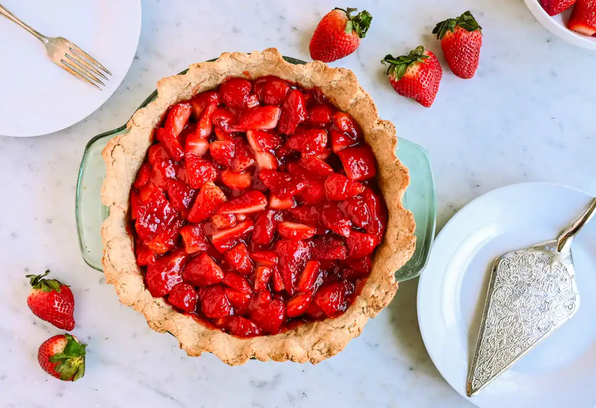 A freshly baked strawberry pie with a golden, flaky sourdough crust, filled with vibrant, juicy strawberries coated in a glossy homemade glaze. The rustic, whole wheat crust adds a rich, nutty flavor that perfectly complements the sweet and tangy strawberry filling. Served in a classic glass pie dish, this delicious dessert is ready to be enjoyed with a fork and plate for the perfect homemade treat.