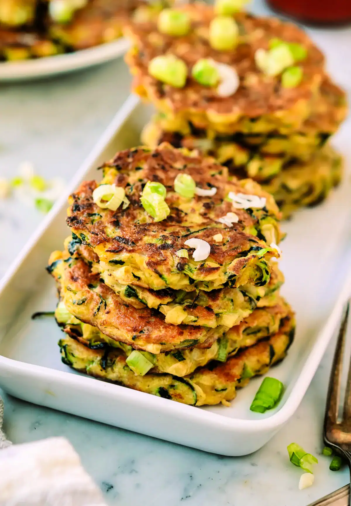 Stacks of homemade zucchini fritters on a white plate, garnished with green onions and served with dipping sauce – easy, kid-friendly sourdough discard recipe.