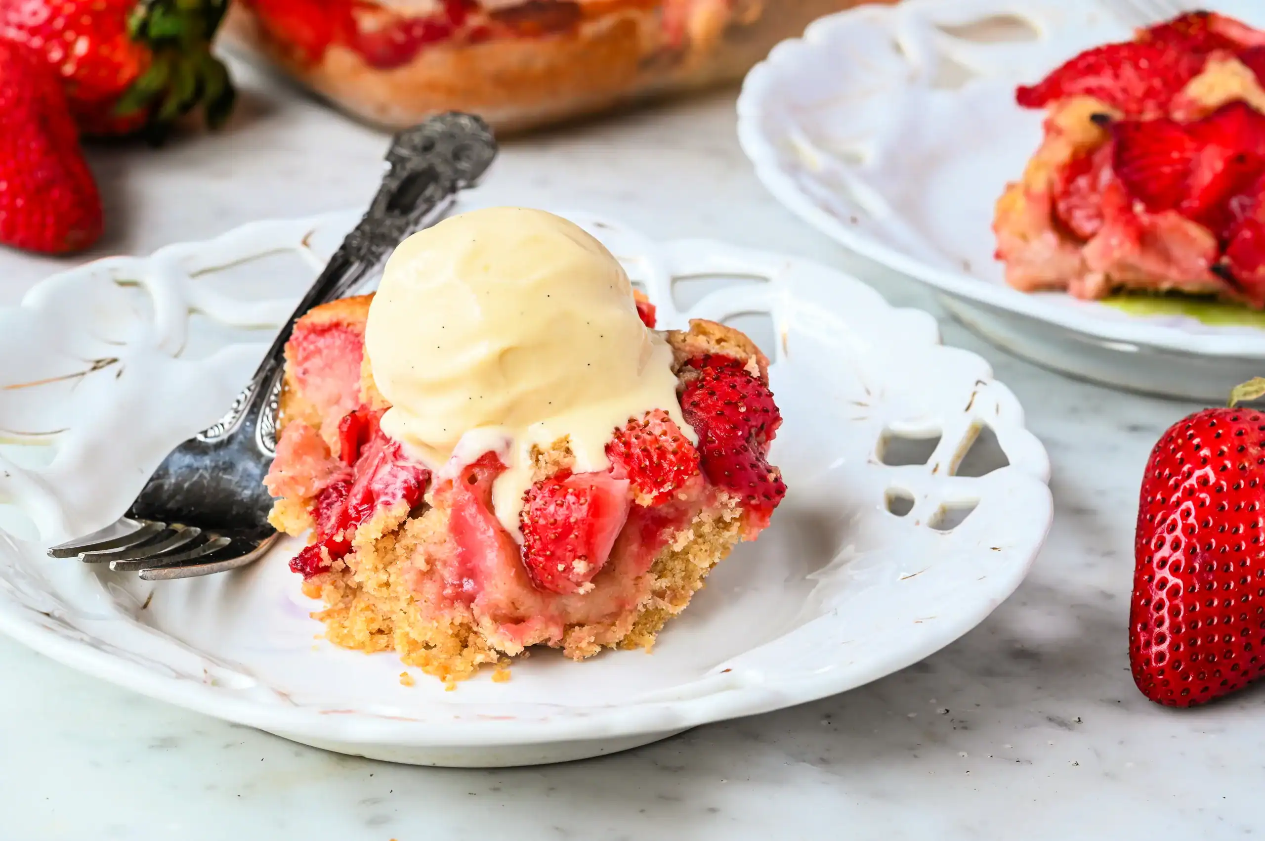 A close-up of a serving of homemade strawberry cobbler with golden, crumbly edges and juicy baked strawberries, topped with a scoop of vanilla ice cream that is beginning to melt. The dessert sits on an elegant white plate with a vintage design, with a silver fork resting beside it. In the background, another plate with a serving of cobbler and fresh strawberries are visible on a marble countertop. The image captures a warm, inviting homemade treat perfect for summer.
