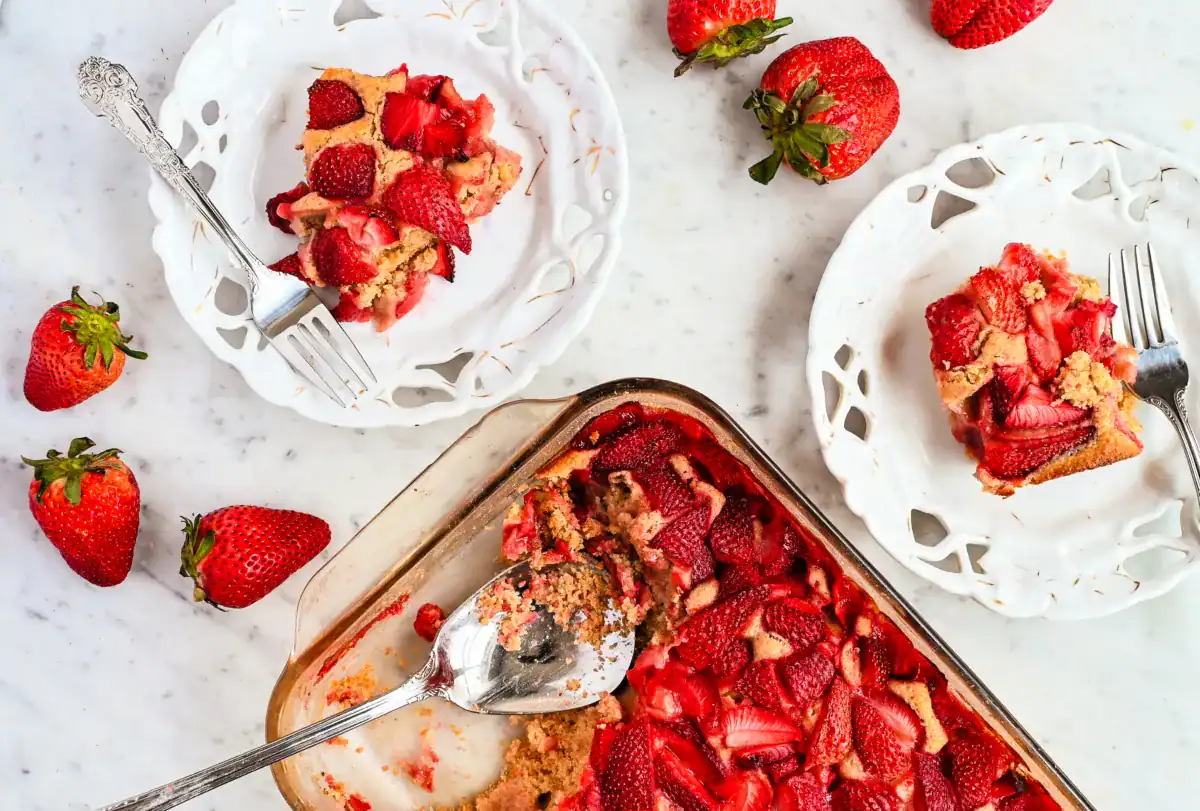 A freshly baked strawberry cobbler in a glass baking dish with a golden, crumbly texture and vibrant red strawberries on top. A large serving spoon rests inside, showing a scoop taken out. Two white vintage-style plates hold individual servings of the cobbler, with silver forks resting beside them. Fresh strawberries are scattered around on the marble countertop, adding to the rustic, homemade feel of the dessert presentation.