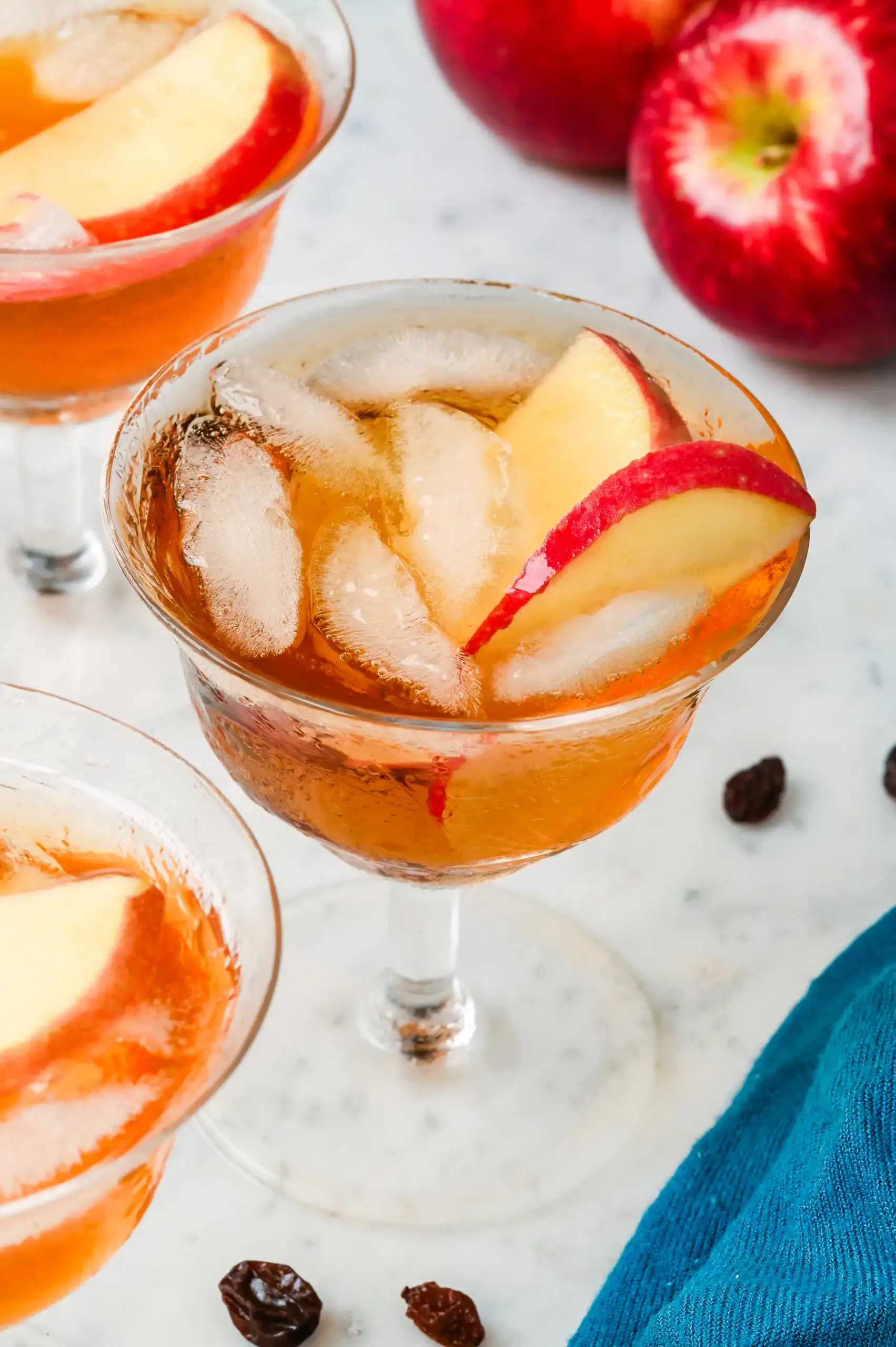 Chilled homemade apple brandy served over ice with fresh apple slices in a glass, surrounded by whole apples and raisins.