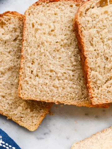 Close-up of sliced sourdough sandwich bread showing soft, airy crumb and golden crust, laid out on a marble surface with a blue kitchen towel.