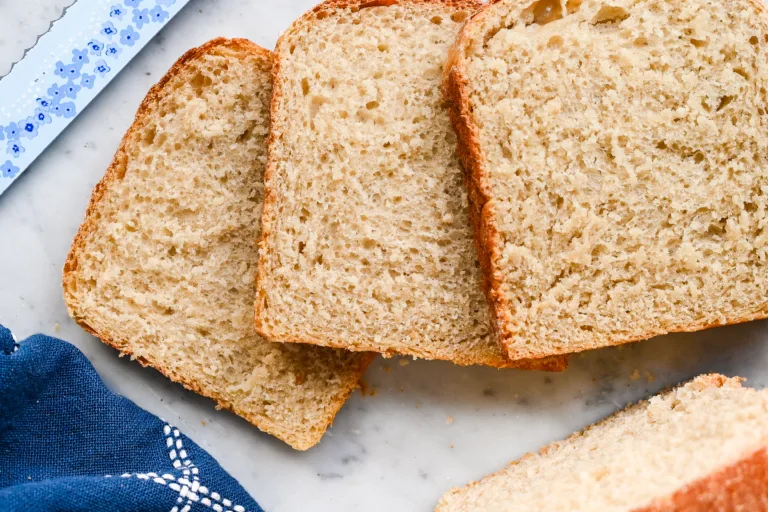 Close-up of sliced sourdough sandwich bread showing soft, airy crumb and golden crust, laid out on a marble surface with a blue kitchen towel.