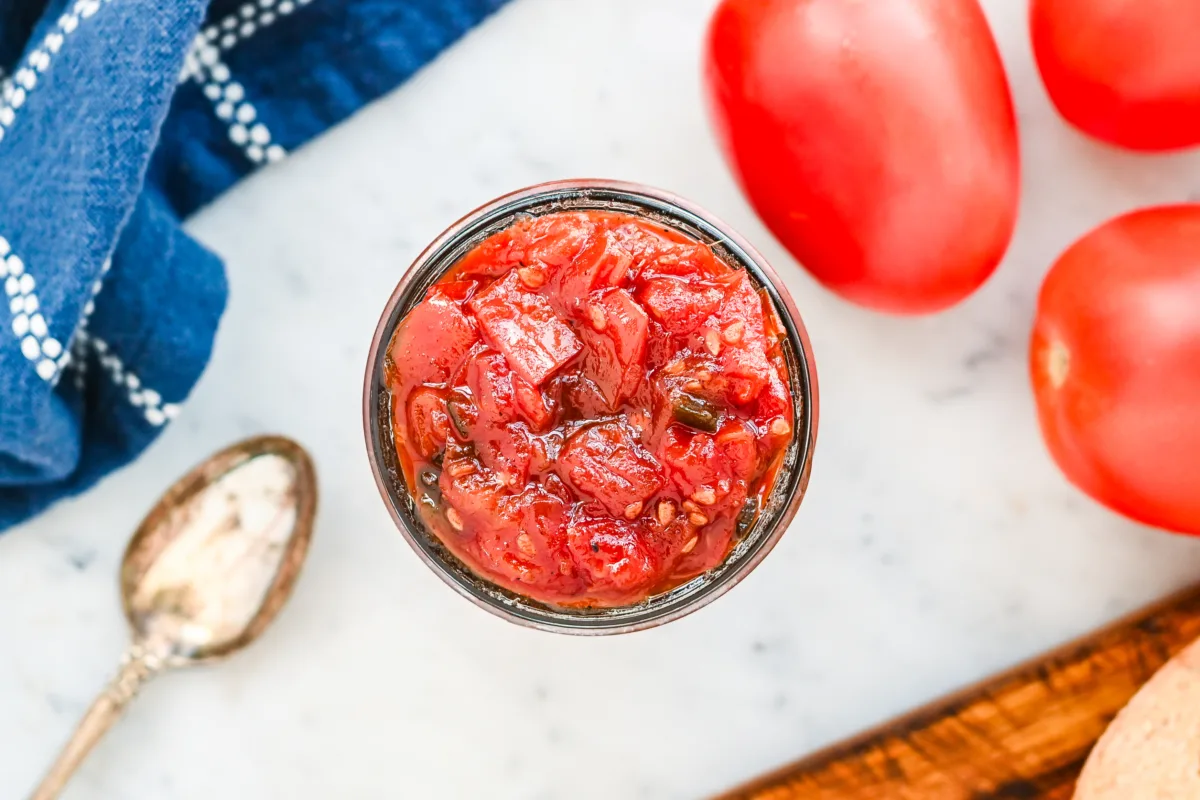 Overhead view of chunky homemade tomato jam in a glass jar, surrounded by fresh tomatoes.