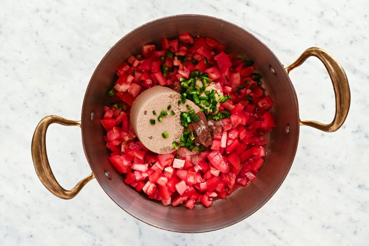 Chopped tomatoes, jalapeños, garlic, and spices in a copper pot with brown sugar ready to make tomato jam.