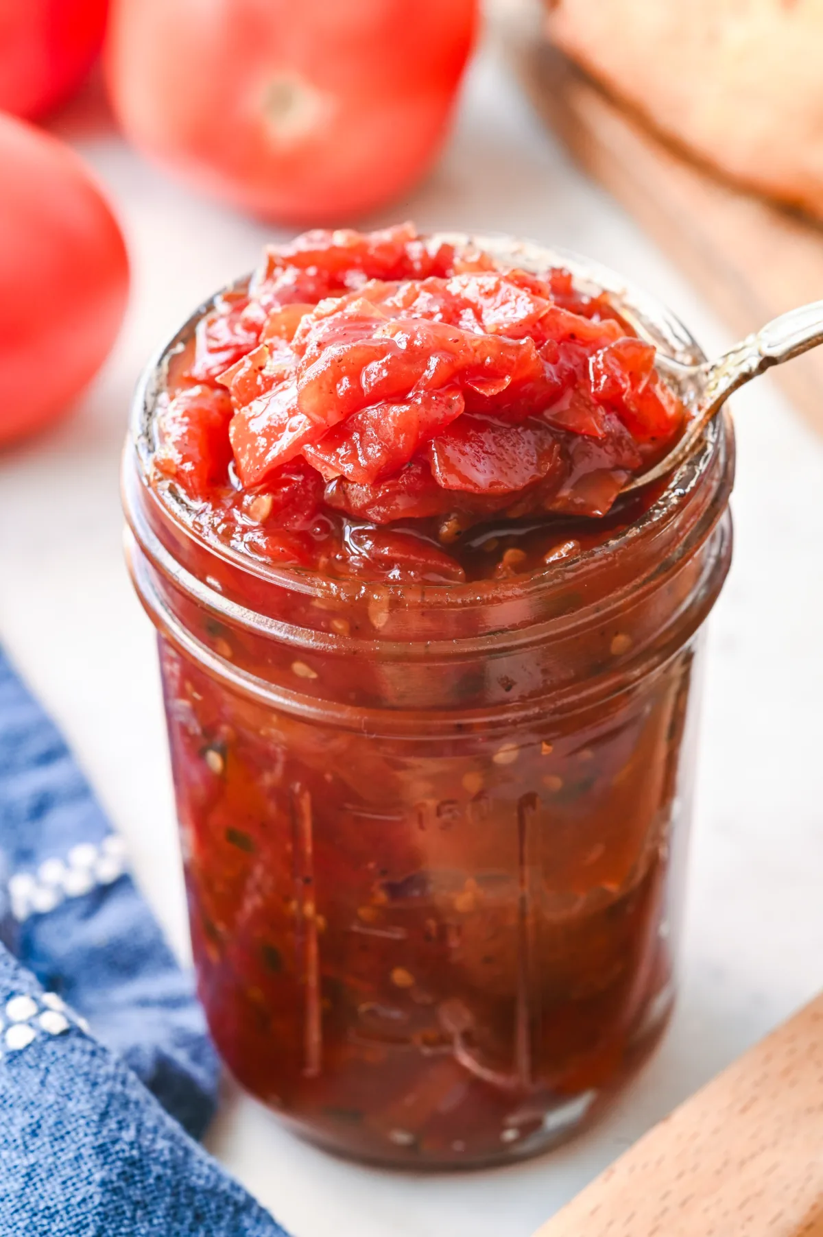 Freshly made tomato jam in a mason jar with ripe tomatoes and bread in the background.