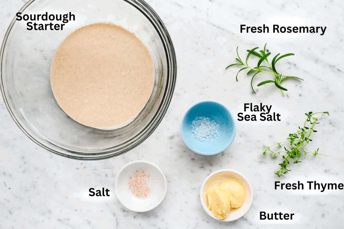Ingredients for thin and crispy sourdough discard crackers, including unfed sourdough starter, sea salt, herbs, and butter on a marble countertop