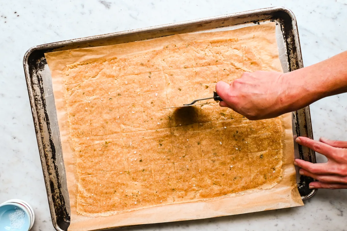 Spreading and trimming thin sourdough cracker dough on a parchment-lined baking sheet—made with unfed sourdough starter and fresh herbs