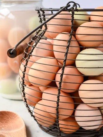 Wire egg basket filled with colorful homestead eggs, with water-glassed eggs in a jar in the background.