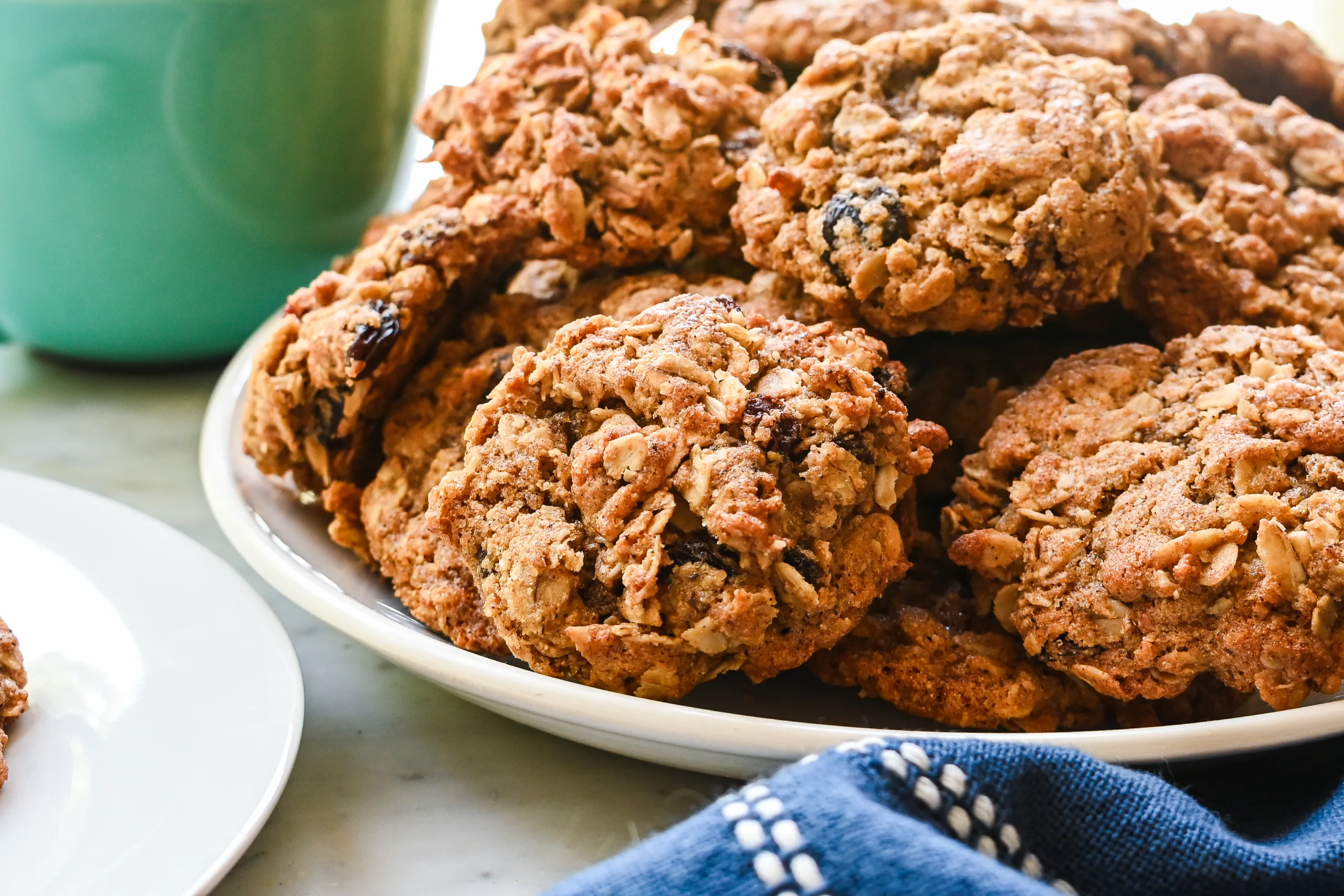 Freshly baked healthy oatmeal raisin cookies stacked on a white plate with a mug in the background.