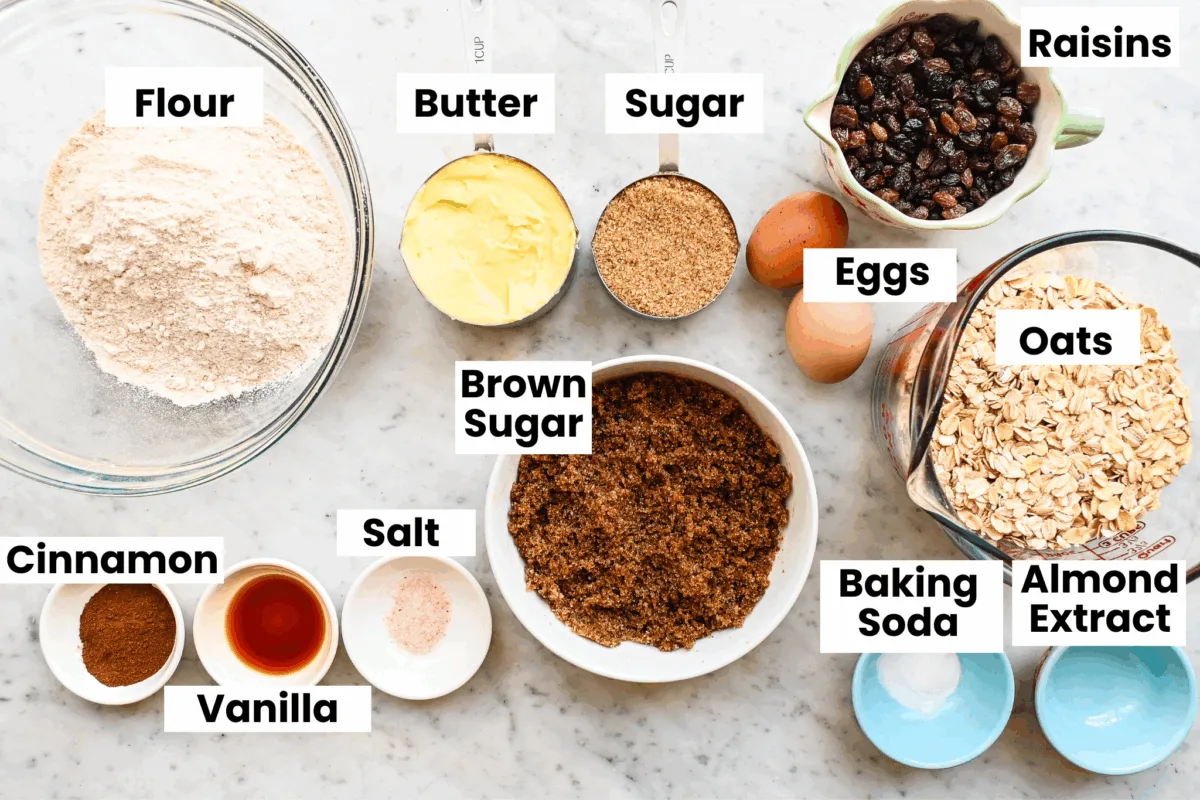 All ingredients for oatmeal raisin cookies arranged on a marble surface, including oats, flour, butter, and raisins.