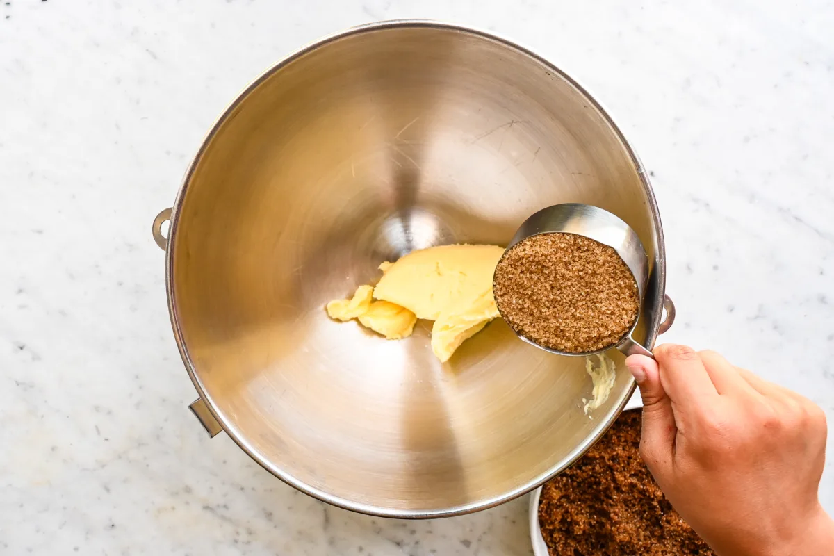 Adding coconut sugar to softened butter in a metal mixing bowl to begin cookie dough preparation.