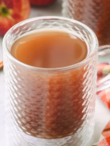 Close-up of a glass mug filled with steaming hot apple cider, surrounded by apples, cinnamon sticks, and a cozy plaid cloth.
