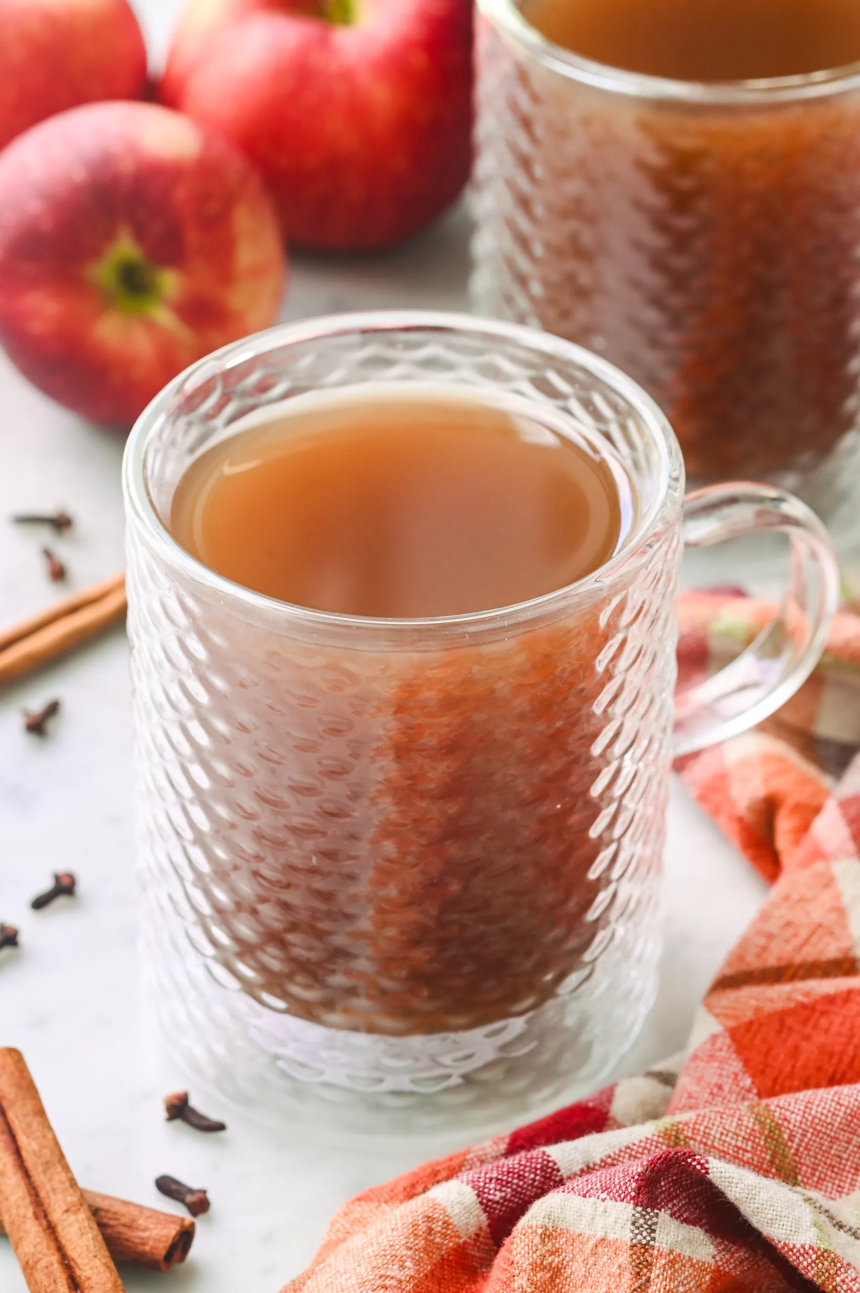 Close-up of a glass mug filled with steaming hot apple cider, surrounded by apples, cinnamon sticks, and a cozy plaid cloth.