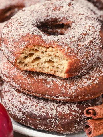 Stack of cinnamon sugar-coated sourdough apple cider donuts, one with a bite showing fluffy interior.
