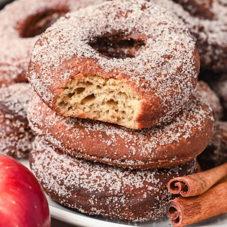 Stack of cinnamon sugar-coated sourdough apple cider donuts, one with a bite showing fluffy interior.