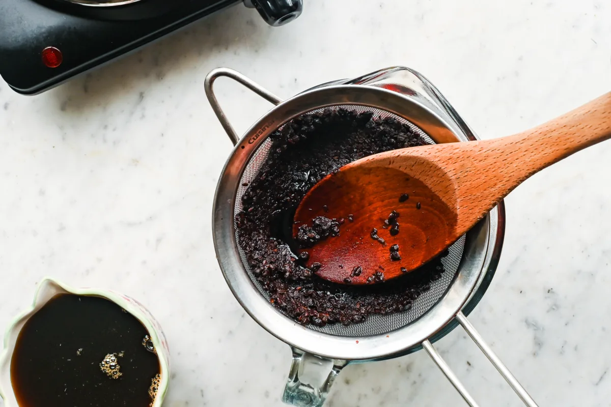 Cooked elderberries and spices being pressed through a strainer with a wooden spoon to separate juice for elderberry syrup.