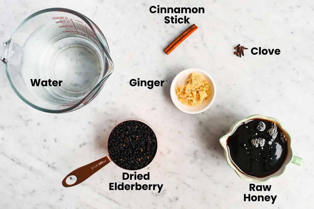 Ingredients for homemade elderberry syrup recipe — dried elderberries, raw honey, ginger, cinnamon stick, cloves, and water measured out on a marble counter.