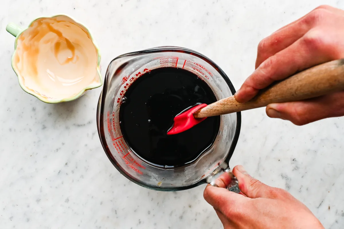 Hands stirring raw honey into strained elderberry juice in a glass measuring cup to finish the syrup.