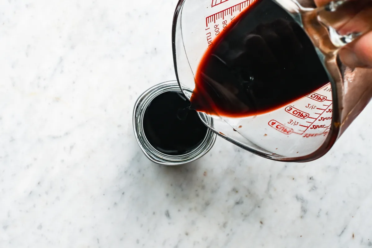 Dark homemade elderberry syrup being poured from a glass measuring cup into a clean glass jar.
