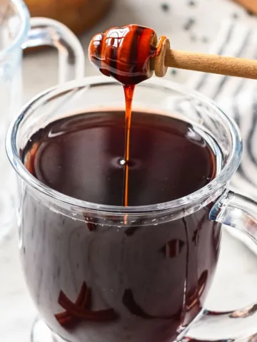 Glass mug of hot elderberry tea sweetened with raw honey, an herbal tea known for its immune-boosting properties, with cinnamon sticks on the side.