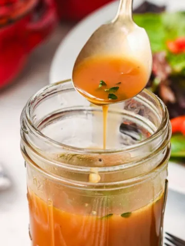 Spoon drizzling homemade salad dressing with fresh herbs from a mason jar over a green salad.