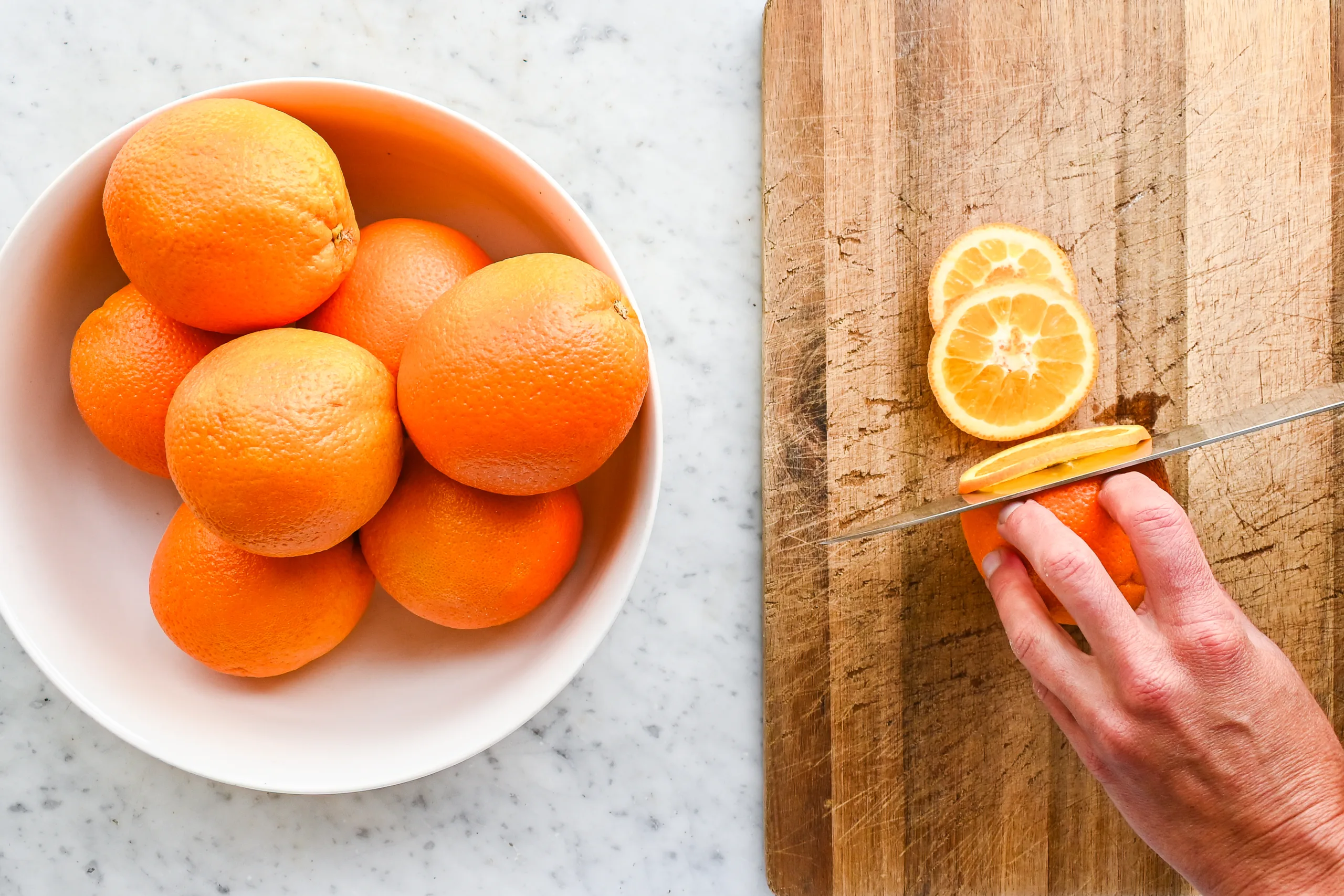 Hand slicing fresh oranges into thin rounds on a wooden cutting board next to a bowl of whole oranges.
