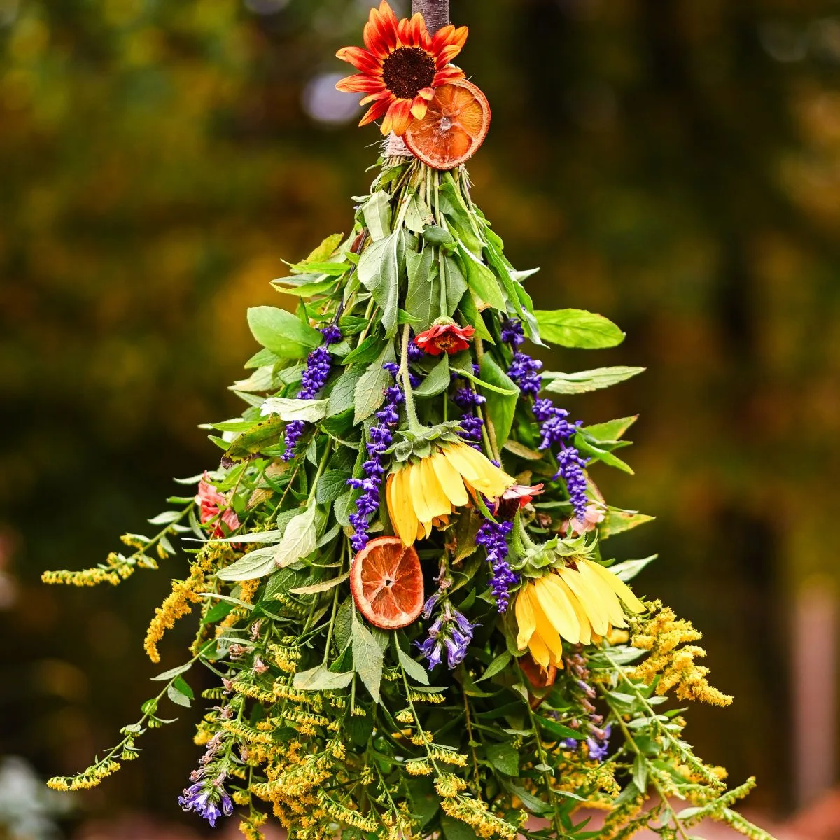 A handmade rustic fall broom with vibrant flowers, greenery, and dried oranges hanging outside against a blurred natural background.