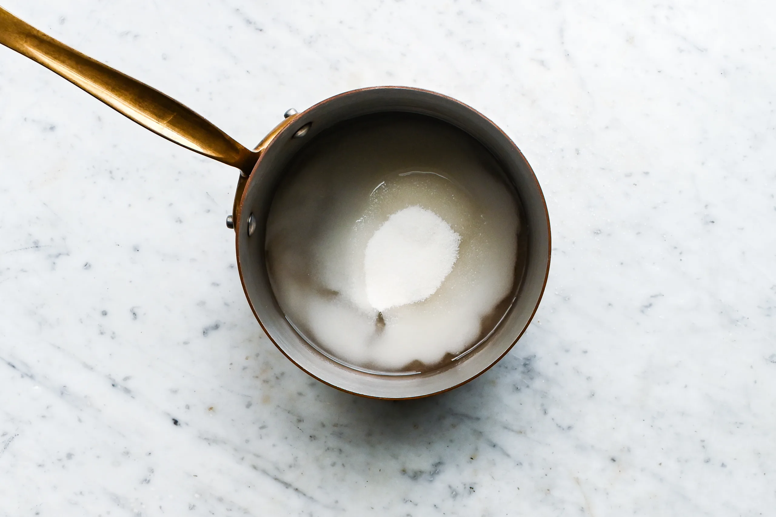 A copper saucepan on marble containing sugar and water before being heated for marshmallow cream.