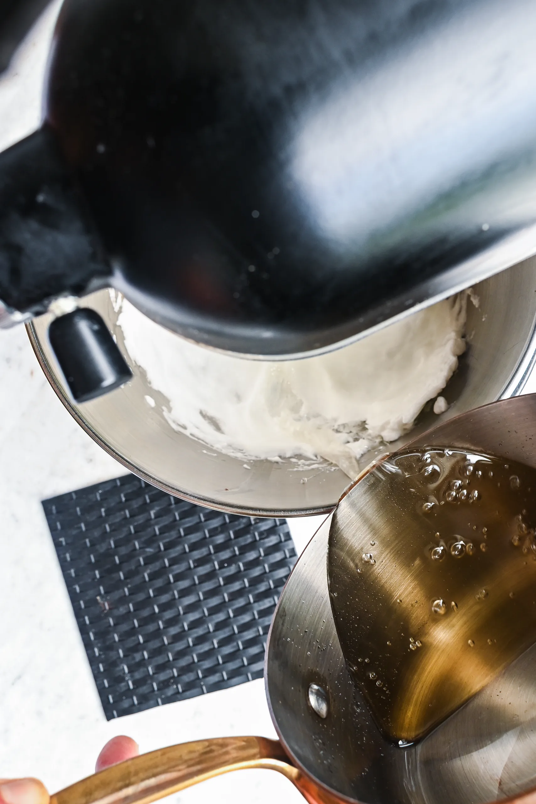 Hot sugar syrup being poured from a saucepan into a stand mixer whipping egg whites.