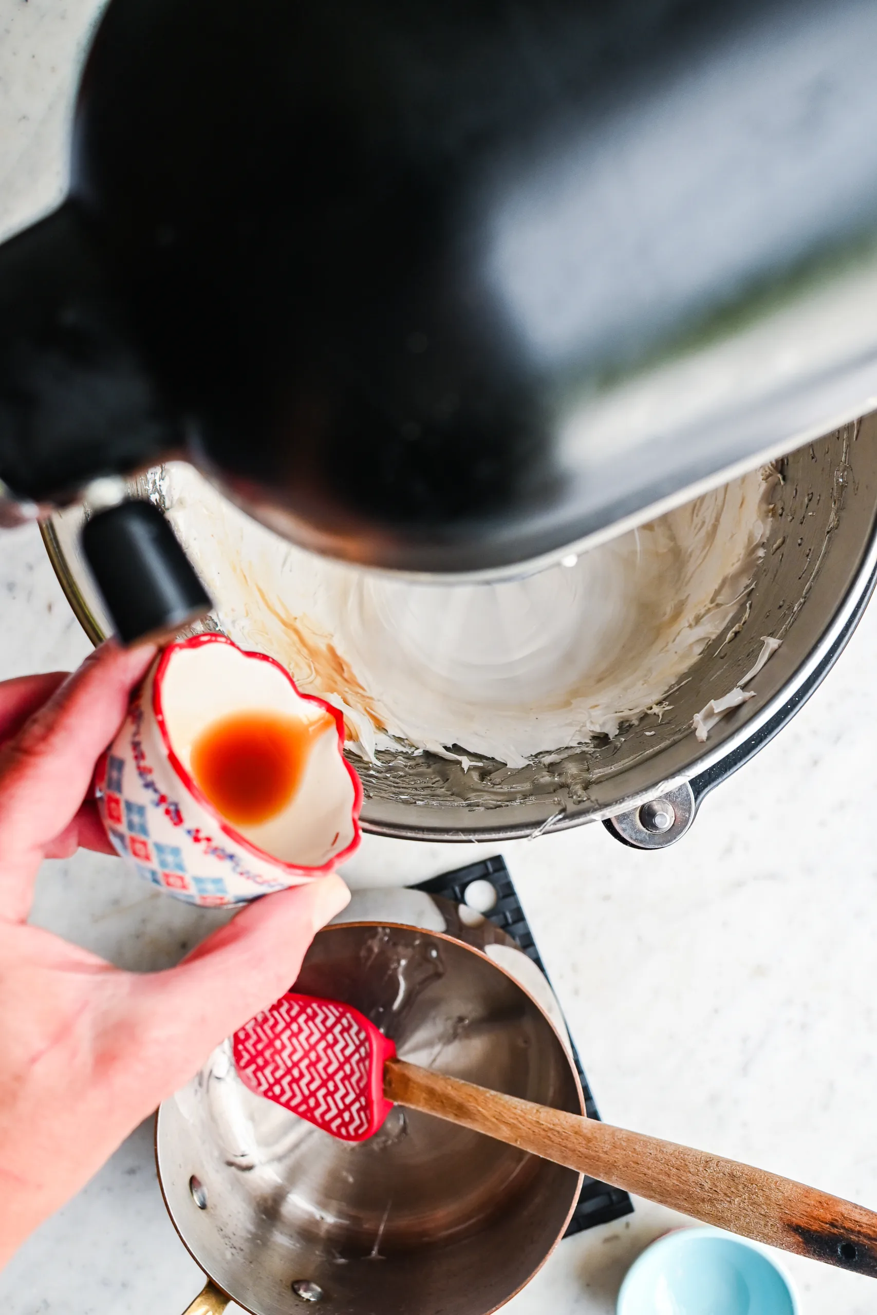 A hand pouring vanilla extract into a stand mixer while fluffy marshmallow cream is being whipped.