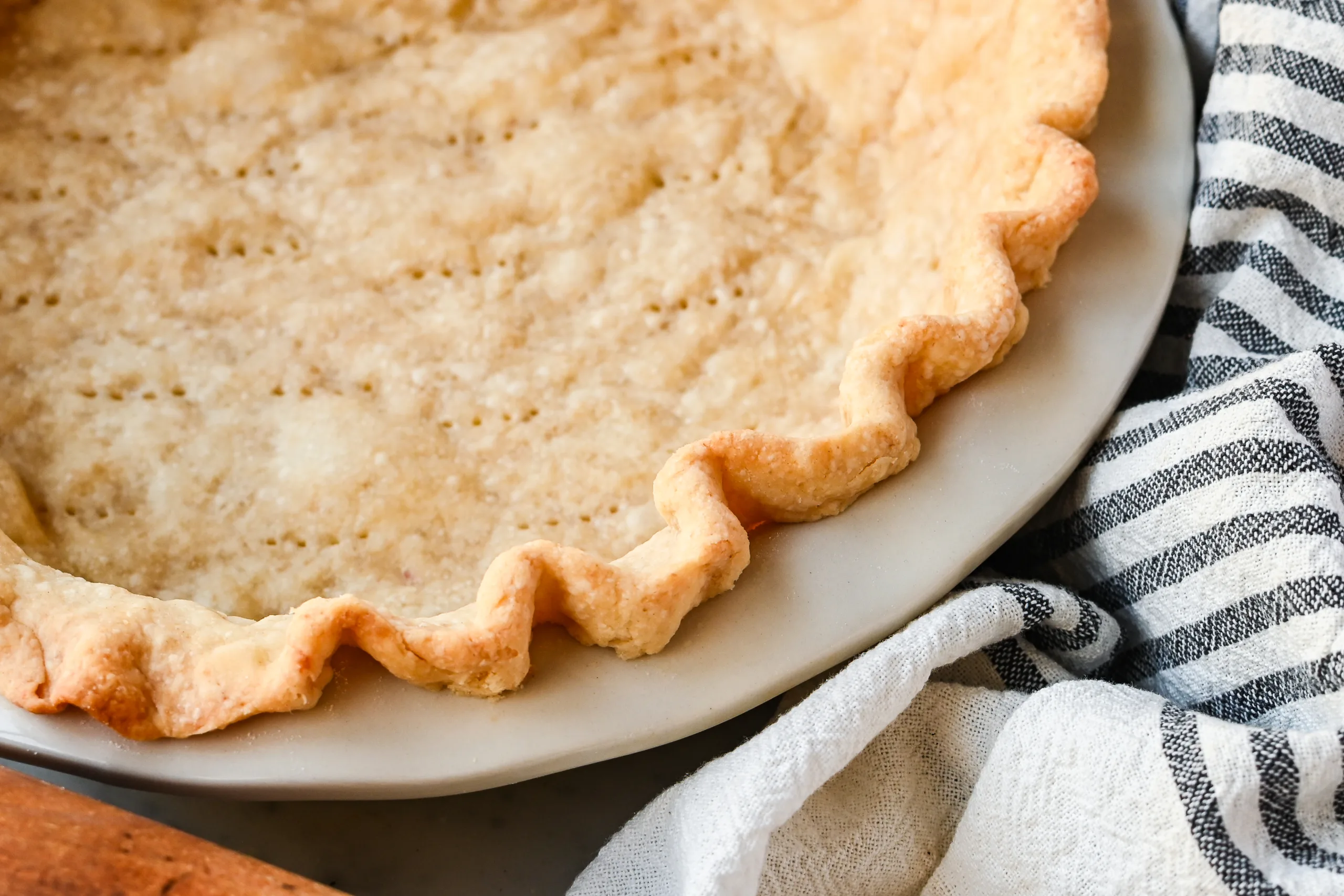 Golden baked sourdough pie crust with crimped edges and fork holes, cooling in a white pie dish on a marble surface with a rolling pin nearby.