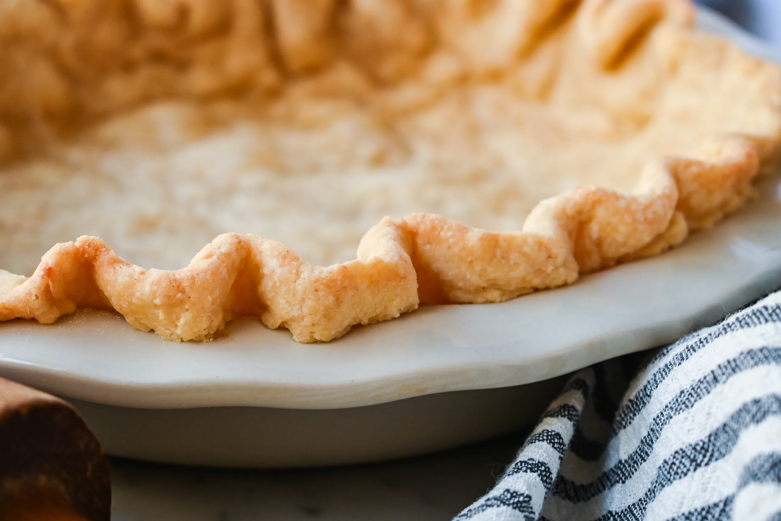 Golden baked sourdough pie crust with crimped edges and fork holes, cooling in a white pie dish on a marble surface with a rolling pin nearby.