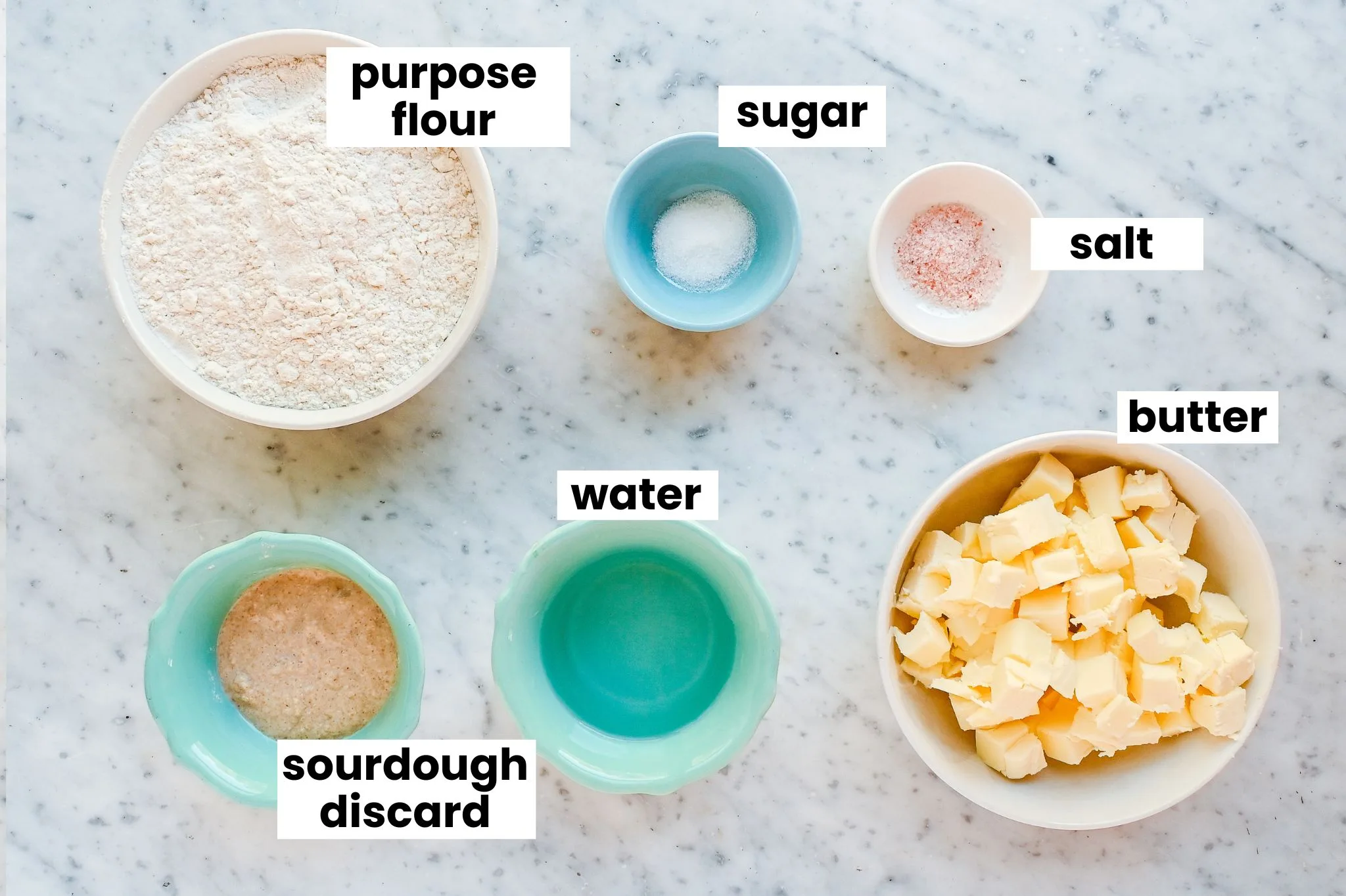 Ingredients for sourdough pie crust measured out in bowls on a marble surface, including flour, butter cubes, sourdough starter, water, and salt.