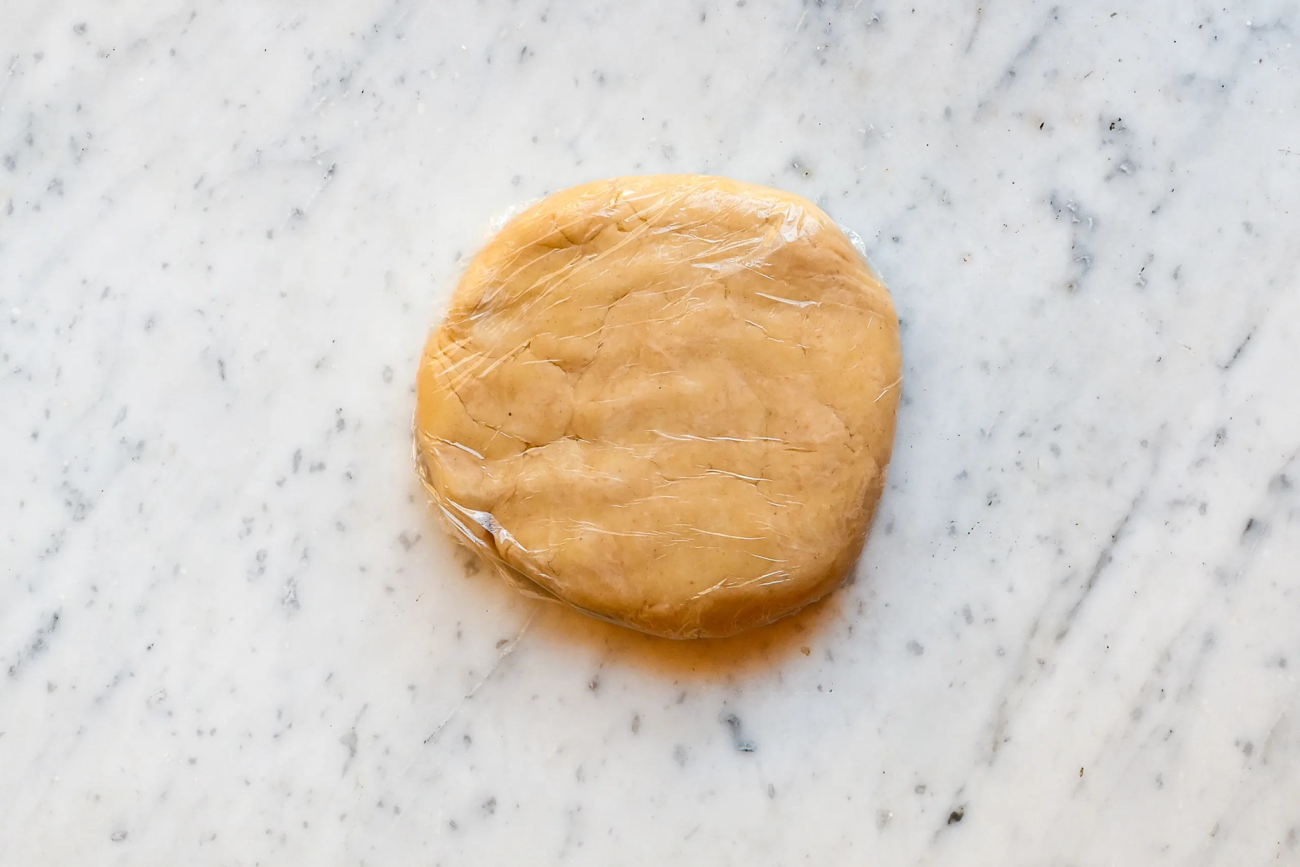Disk of sourdough pie dough wrapped in plastic wrap resting on a marble surface, ready to chill before rolling out.