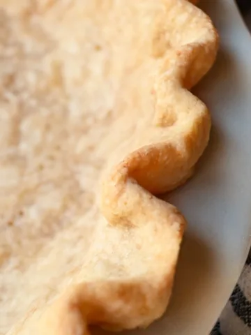 Golden baked sourdough pie crust with crimped edges and fork holes, cooling in a white pie dish on a marble surface with a rolling pin nearby.