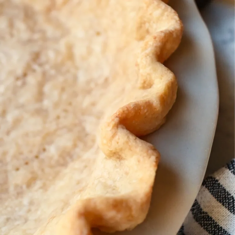 Golden baked sourdough pie crust with crimped edges and fork holes, cooling in a white pie dish on a marble surface with a rolling pin nearby.