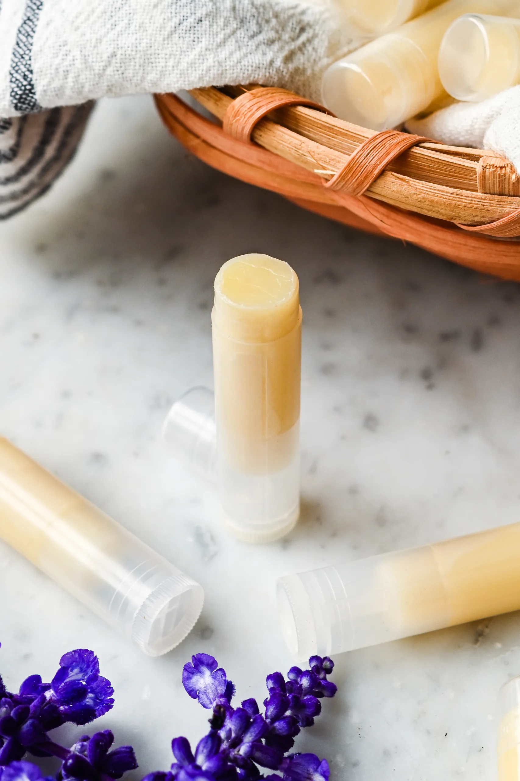 Finished homemade tallow lip balm tubes on a marble countertop, with a basket and purple flowers in the background.