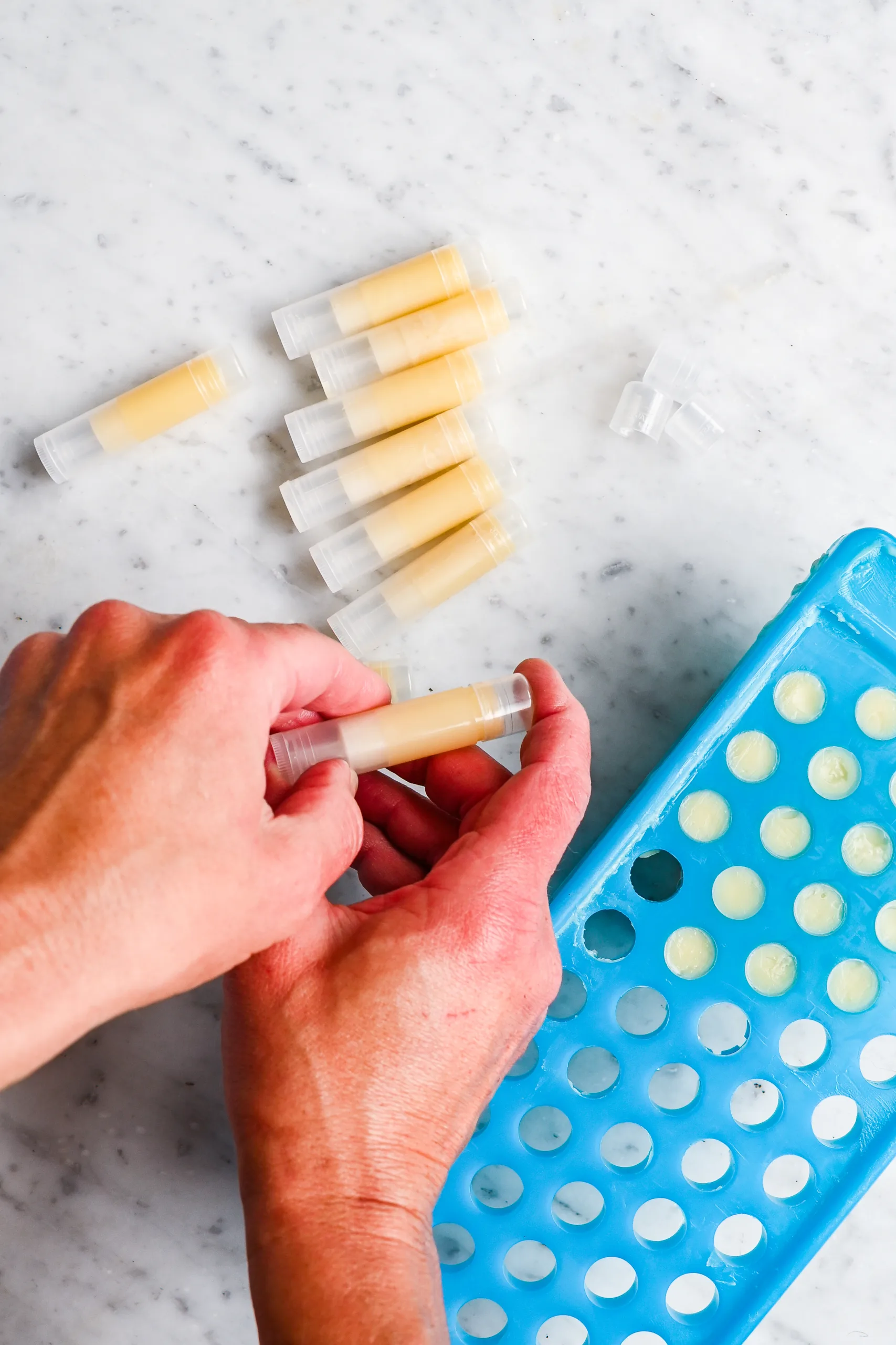 Hands placing homemade lip balm into tubes using a blue silicone mold on a marble surface, with several filled tubes lying nearby.
