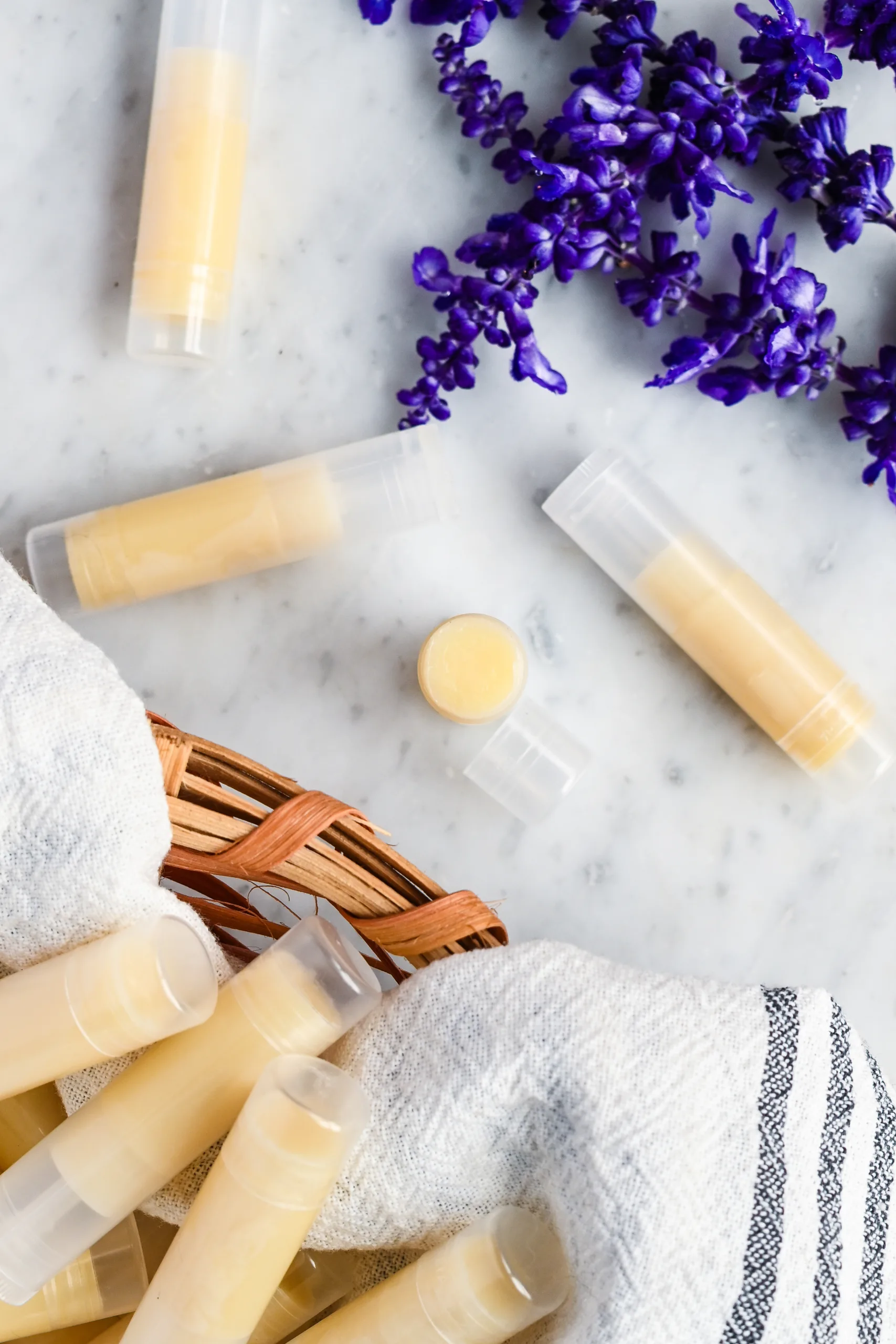 Finished homemade tallow lip balm tubes on a marble countertop, with a basket and purple flowers in the background.