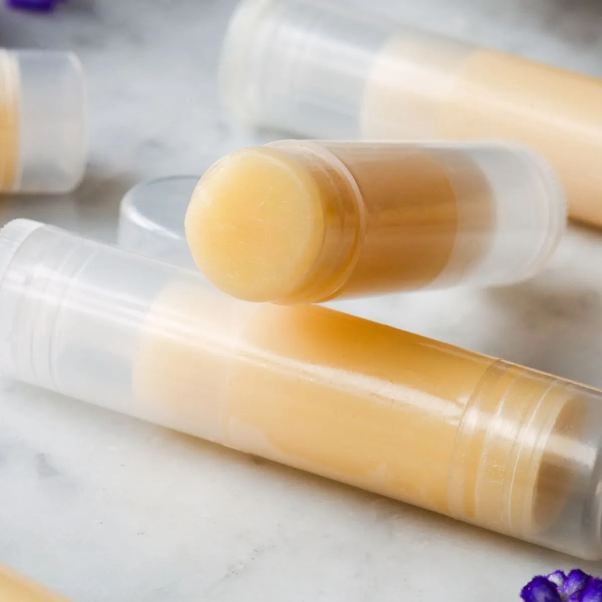 Finished homemade tallow lip balm tubes on a marble countertop, with a basket and purple flowers in the background.