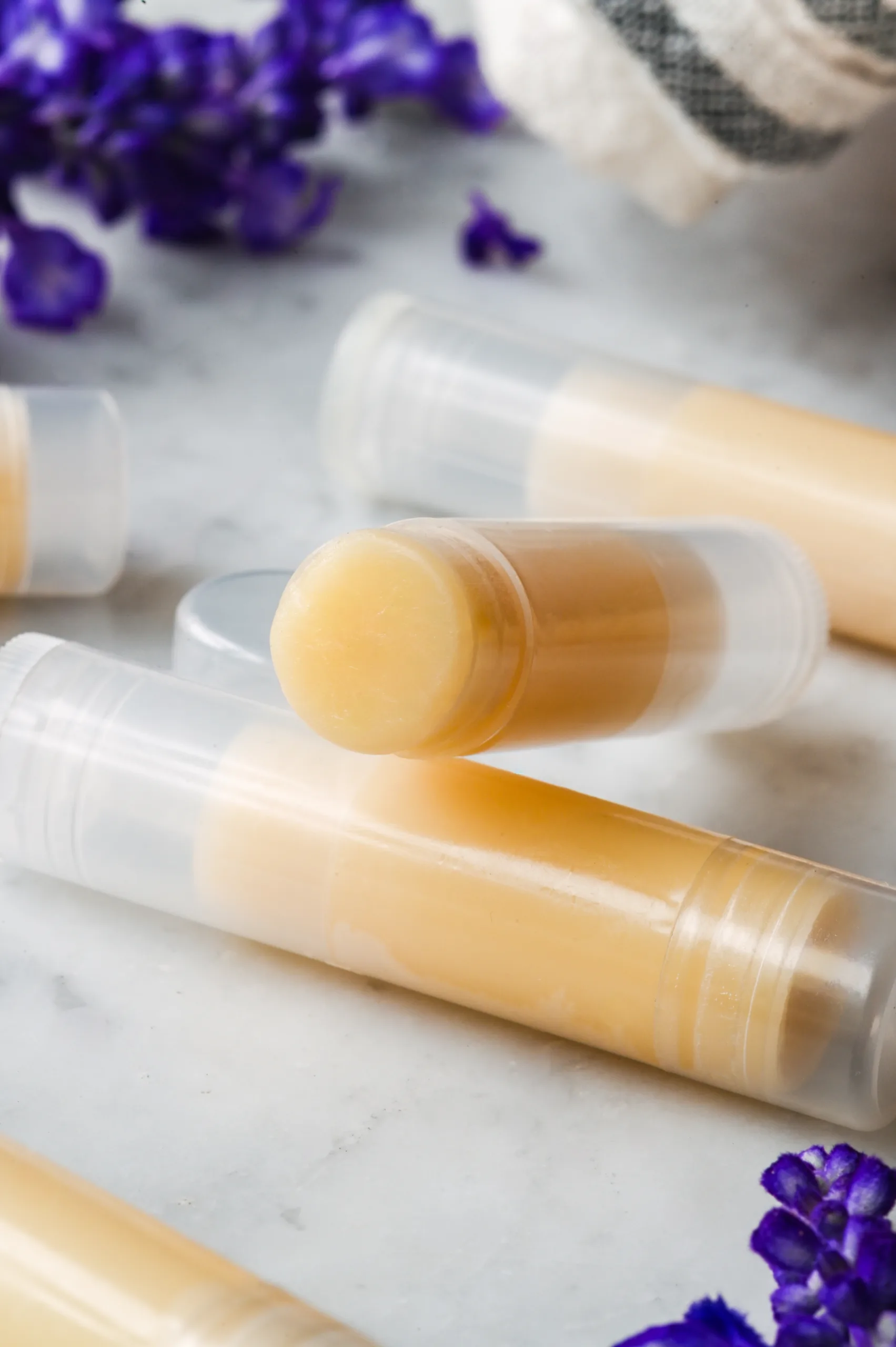 Finished homemade tallow lip balm tubes on a marble countertop, with a basket and purple flowers in the background.
