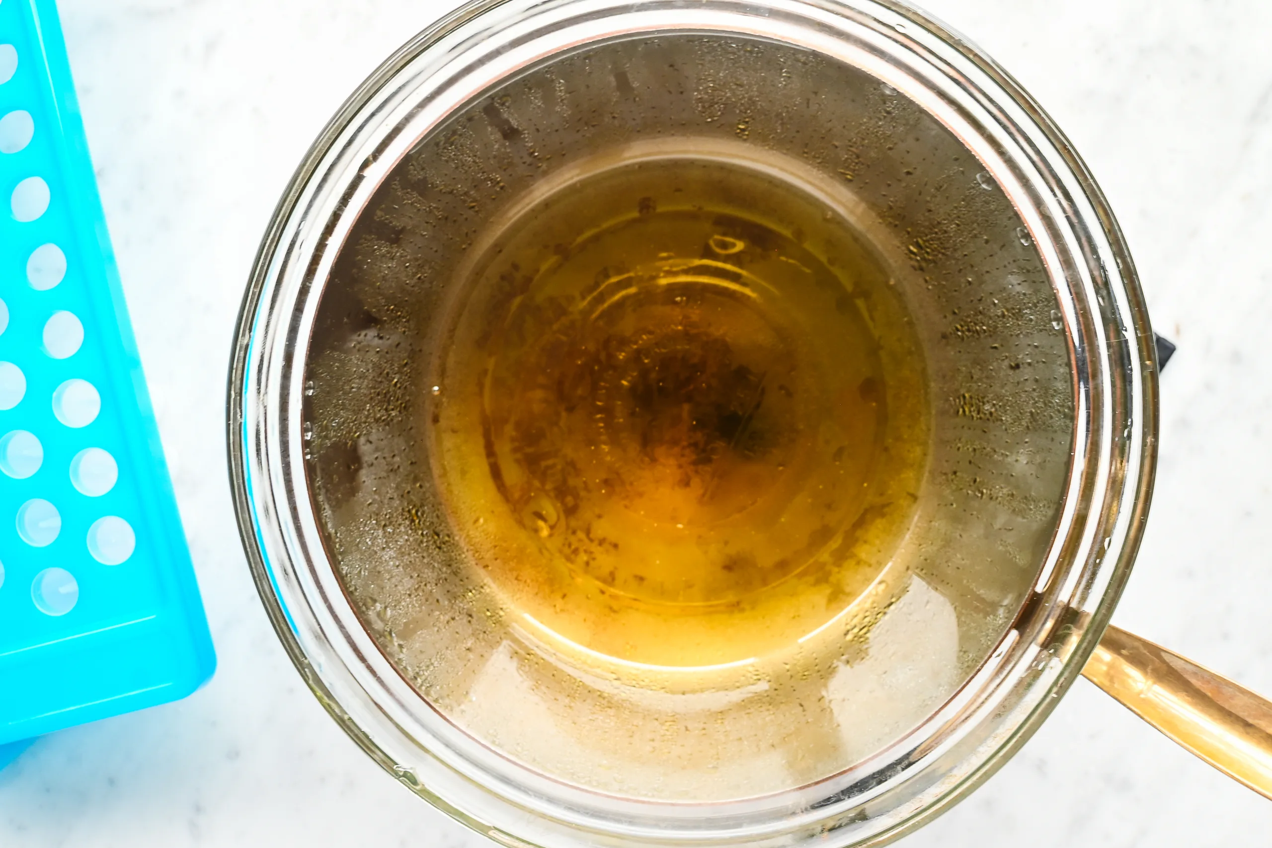 Melted tallow, beeswax, and oils in a glass double boiler bowl, fully liquified and ready to be poured into lip balm molds. A blue silicone mold sits to the side on a marble surface.