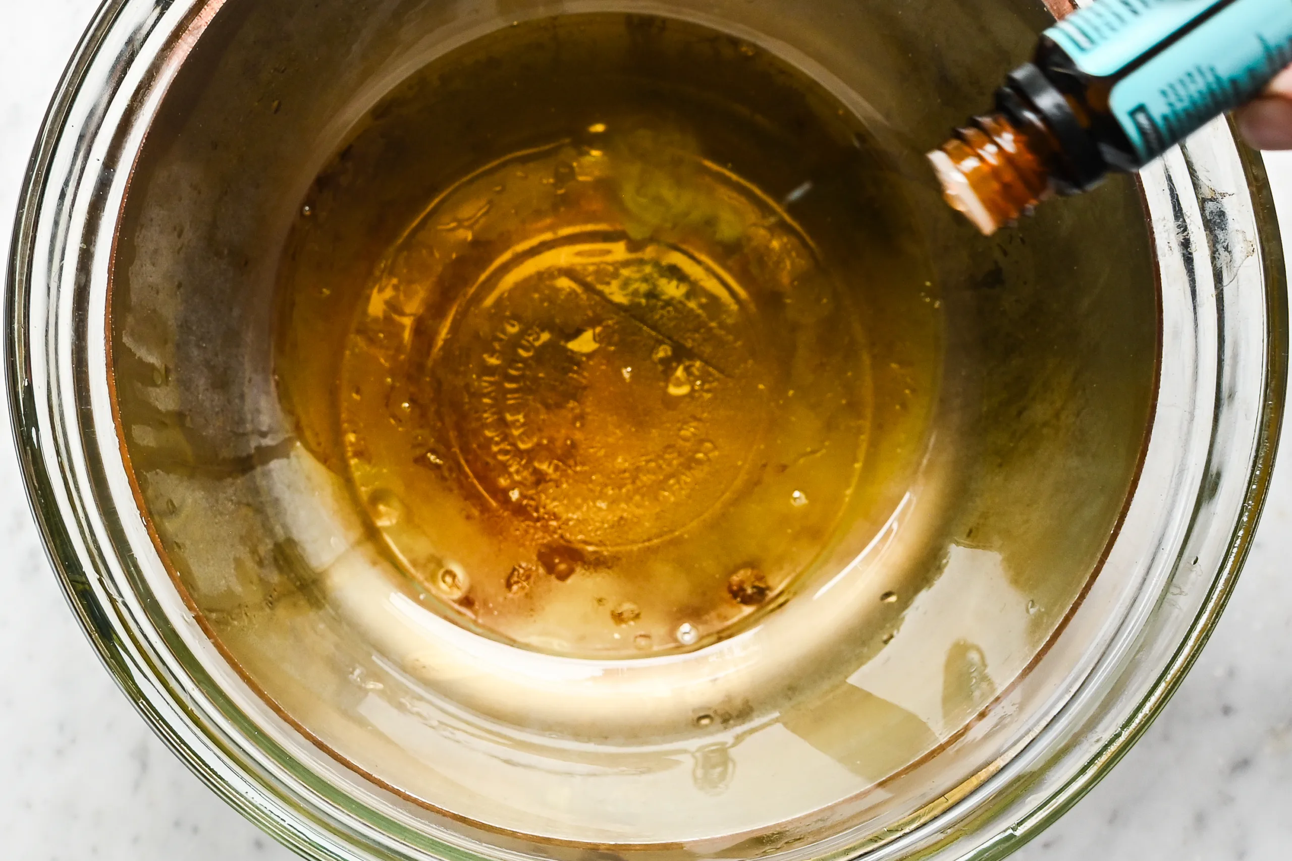 Essential oil being added to the fully melted lip balm mixture in a glass bowl over a double boiler.