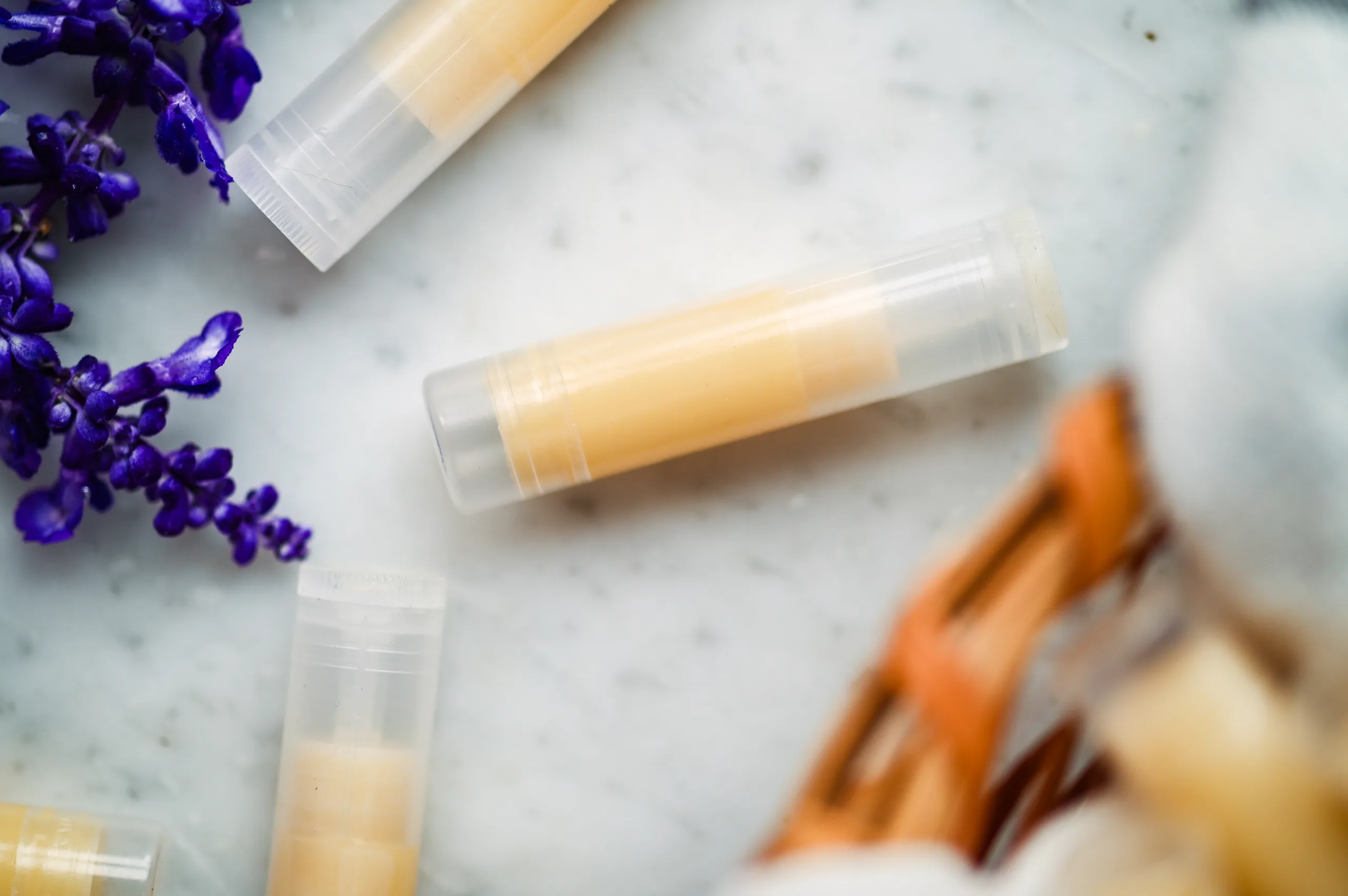Finished homemade tallow lip balm tubes on a marble countertop, with a basket and purple flowers in the background.