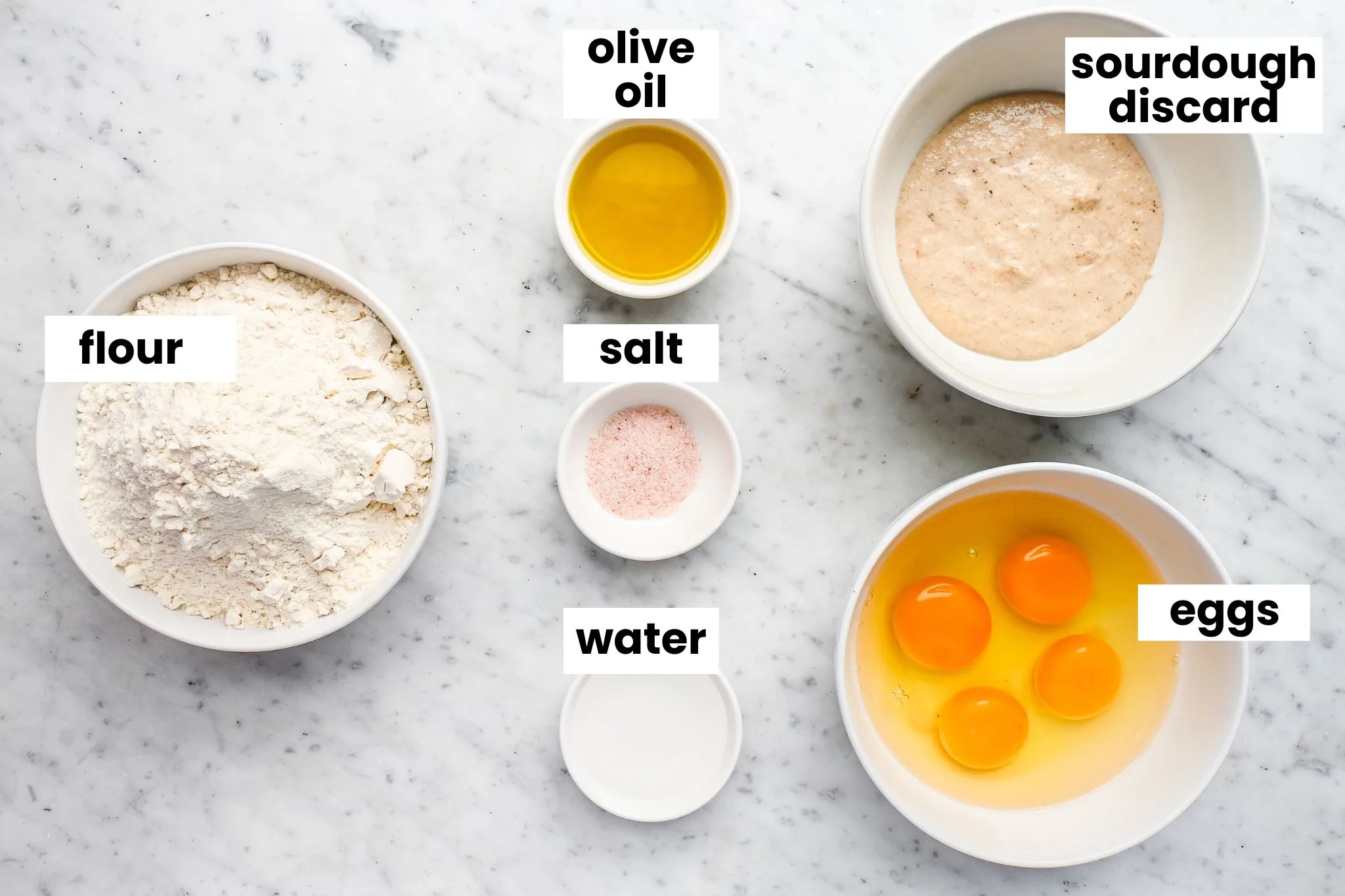 Bowls of flour, eggs, olive oil, sourdough starter discard, salt, and water arranged neatly on a marble surface for making fresh sourdough pasta dough.