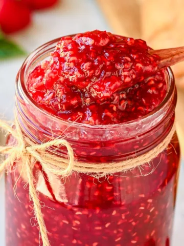 Wooden spoon scooping homemade raspberry jam from a glass jar tied with twine, showing the thick, chunky texture.