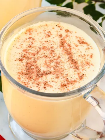 Glass mugs of non-alcoholic eggnog topped with cinnamon and nutmeg, garnished with holly and cinnamon sticks for Christmas.